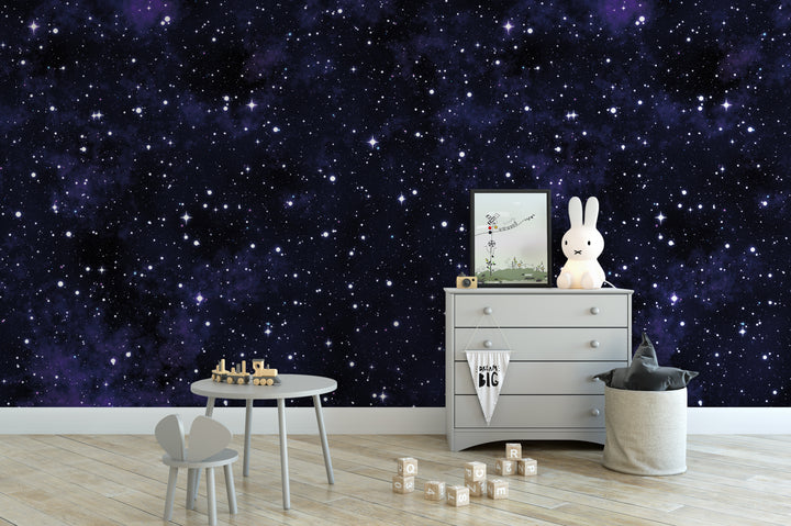 a cozy nursery or child's room with a starry night sky mural on the wall, a wooden table with a toy train, a dresser with a picture frame, and a stuffed bunny toy.
