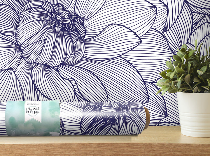 a large blue and white floral pattern on a wall, with a potted plant and a roll of wallpaper or paper with a floral design on a shelf in the foreground.