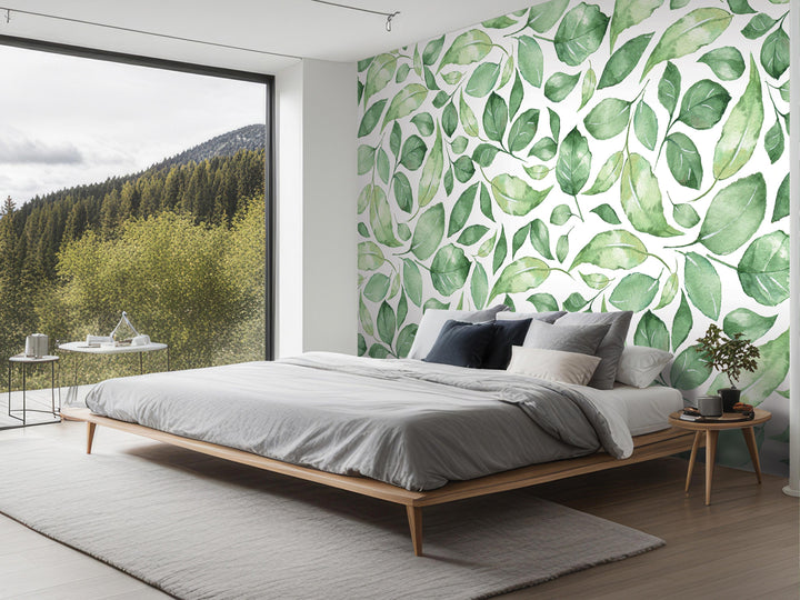 a modern bedroom with a large window overlooking a forested area. The room features a bed with a gray comforter and pillows, a nightstand, and a rug. The wall behind the bed has a large mural of green leaves, creating a natural and serene atmosphere in the space.