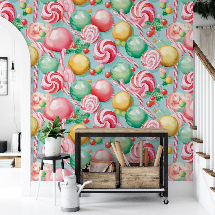 a wallpapered wall with a vibrant and colorful candy-themed design, including various types of candies such as lollipops, jelly beans, and other sweets. In front of the wall, there is a small table with a plant and a white watering can, as well as a wooden crate with some items inside.