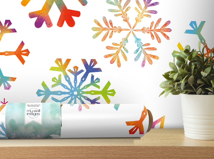 a white wall with a vibrant, colorful snowflake pattern. In the foreground, there is a roll of wallpaper with a snowflake design, and a potted plant is visible to the right of the wallpaper.
