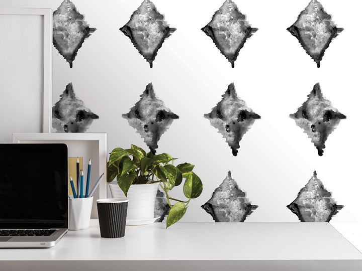 a white desk with a laptop, a plant, and various office supplies such as a cup and a pencil holder. The wall behind the desk has a repeating pattern of black and white abstract shapes, creating a visually striking backdrop for the desk setup.