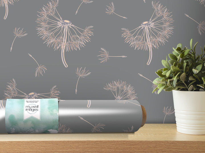 a gray wall with a repeating pattern of dandelion-like designs in shades of pink and white. A roll of wallpaper with a similar pattern is placed on a wooden shelf next to a potted plant.