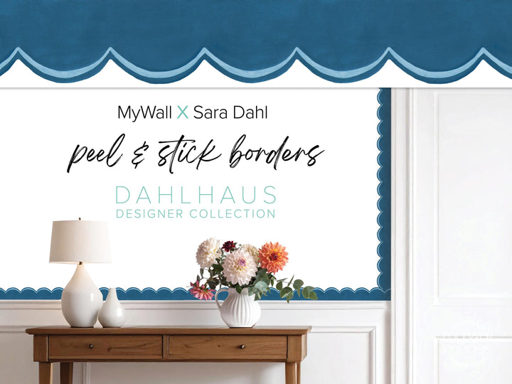 a wooden table with a vase of flowers, a lamp, and a blue and white patterned curtain. The curtain has a scalloped edge and the text "MyWall x Sara Dahl peel & stick borders" is displayed on it.