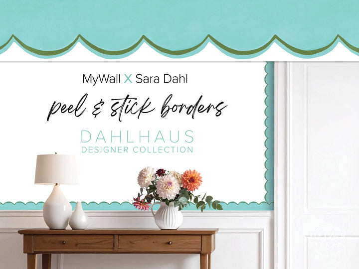 The image is an advertisement for MyWall x Sara Dahl peel and stick borders. It features a wooden table with a vase of flowers, a lamp, and a blue and white scalloped trim. The text on the wall reads "MyWall x Sara Dahl peel & stick borders" and "Dahlhaus Designer Collection".