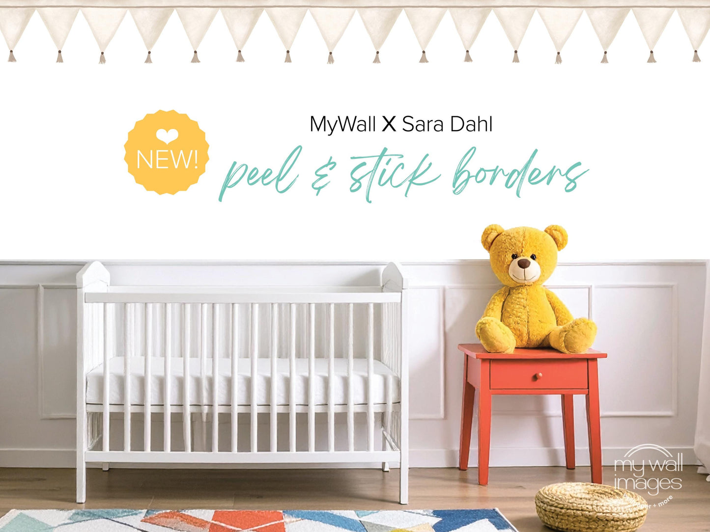 a nursery room with a white crib, a yellow teddy bear, and a red table. The crib is positioned against a white wall, and the teddy bear is sitting on the table. The room has a colorful rug on the floor, and there are white curtains on the windows. The image also includes text that reads "MyWall X Sara Dahl peel & stick borders" and "NEW!"