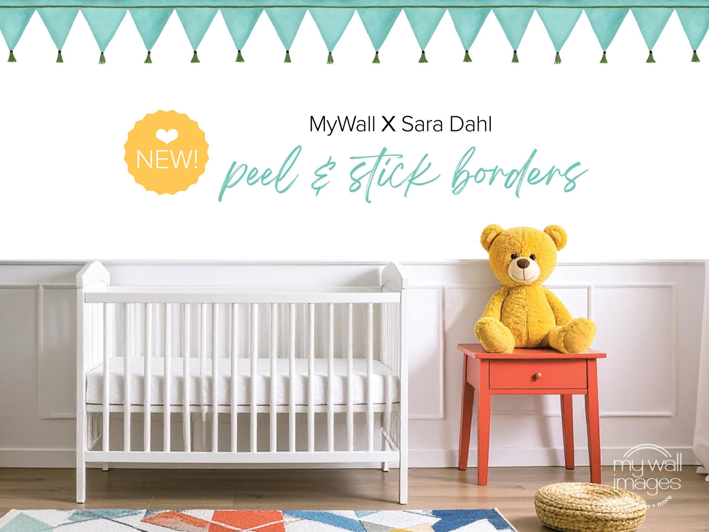 a nursery room with a white crib, a yellow teddy bear, and a red table. The crib is positioned against a white wall, and the teddy bear is sitting on the table. The room has a colorful rug on the floor, and there are green and white tassels hanging from the ceiling. The image includes text that reads "MyWall X Sara Dahl peel & stick borders" and "NEW! peel & stick borders", suggesting that the nursery is being advertised or updated.
