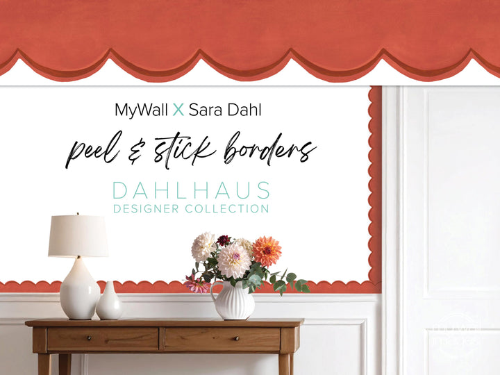 a wooden table with a vase of flowers, a lamp, and a red scalloped trim on the wall behind it. The text on the wall reads "MyWall x Sara Dahl peel & stick borders" and "Dahlhaus Designer Collection".
