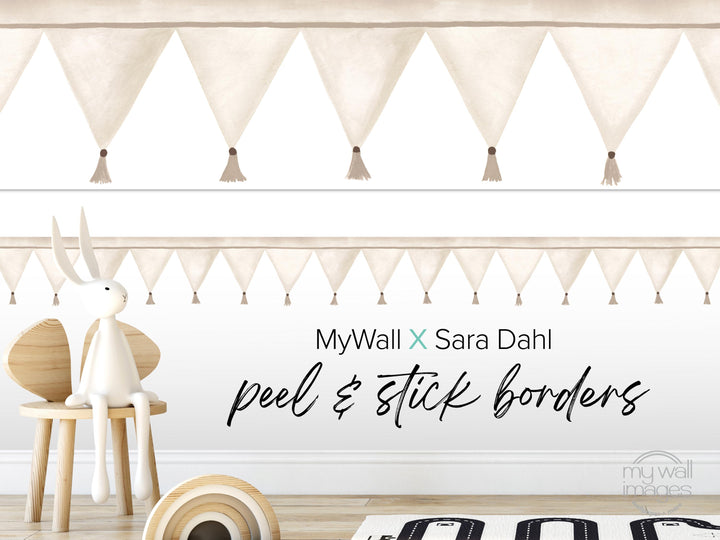 a white rabbit sitting on a wooden chair, with a banner of white triangles hanging on the wall behind it. The banner has tassels hanging from the edges. The text "MyWall X Sara Dahl peel & stick borders" is written on the wall, and there is a small wooden toy rabbit on the floor next to the chair.