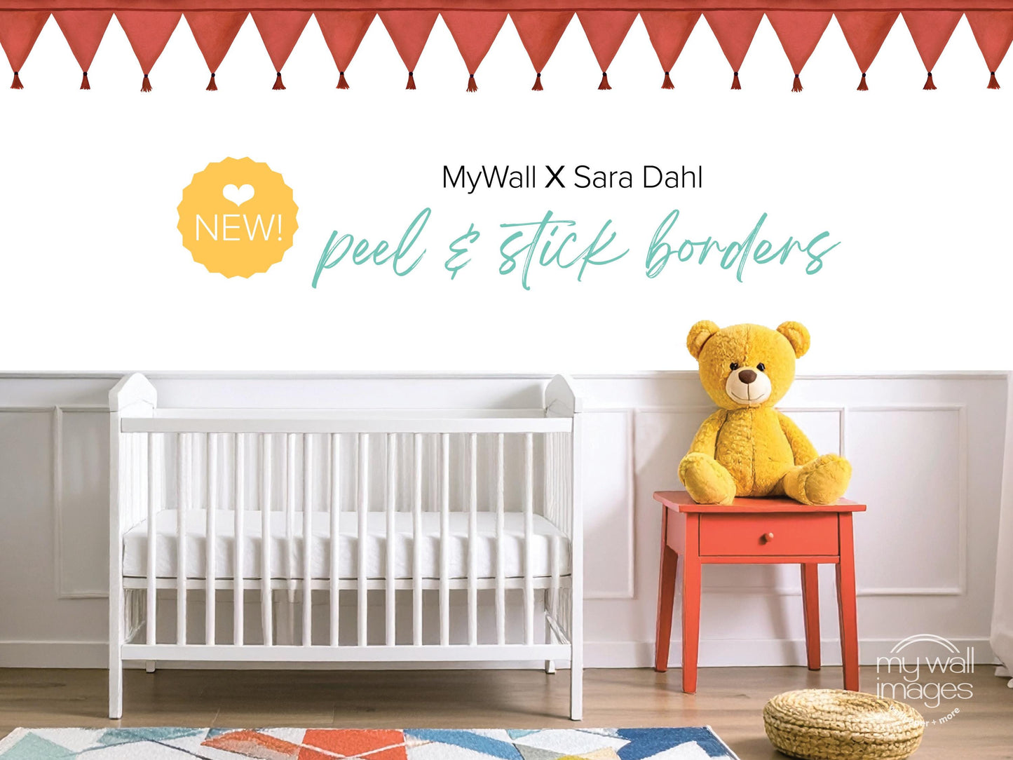 a nursery room with a white crib, a yellow teddy bear, and a red table with a drawer. The crib is positioned against a white wall, and the teddy bear is sitting on the table. The room has a colorful rug on the floor, and there are red and white tassels hanging from the ceiling. The image includes text that reads "MyWall X Sara Dahl peel & stick borders" and "NEW! peel & stick borders", suggesting that the nursery is being advertised or updated with new products.