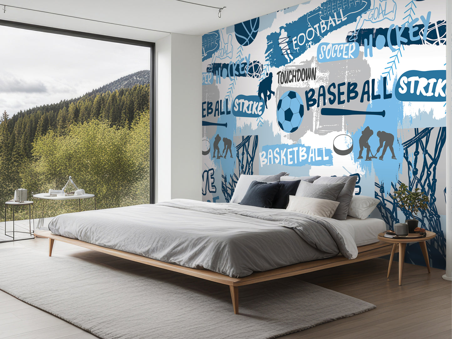 a bedroom with a large wall mural depicting various sports, including football, soccer, baseball, and tennis. The mural is colorful and covers a significant portion of the wall. The room has a bed with a gray comforter and pillows, and there is a small table next to the bed. The room also has a large window overlooking a scenic outdoor view of a forest.