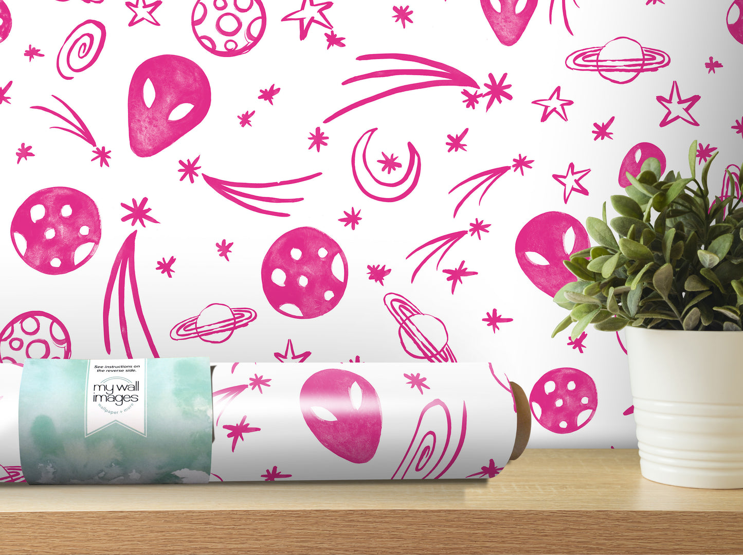 a wallpapered wall with a repeating pattern of pink alien and celestial motifs, including planets, stars, and comets. In the foreground, there is a potted plant on a shelf, and a roll of wallpaper is visible on the shelf as well.