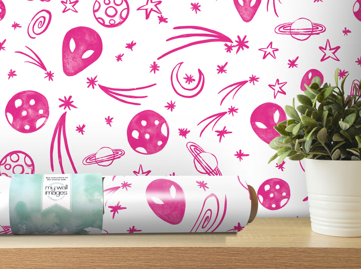 a wallpapered wall with a repeating pattern of pink alien and celestial motifs, including planets, stars, and comets. In the foreground, there is a potted plant on a shelf, and a roll of wallpaper is visible on the shelf as well.