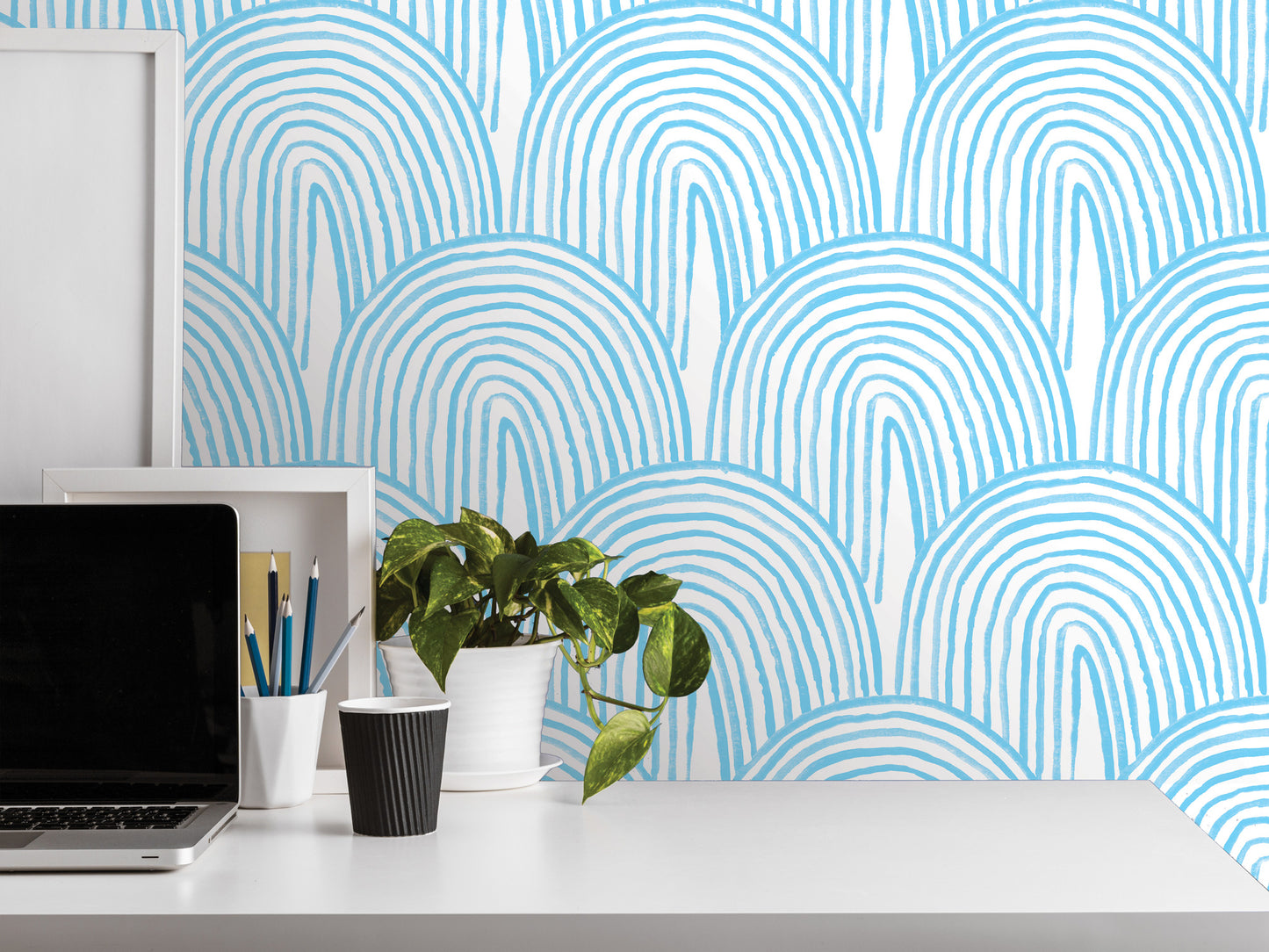 a white desk with a laptop, a black cup, a white vase with a plant, and a framed picture. The desk is set against a wall with a blue and white striped pattern.