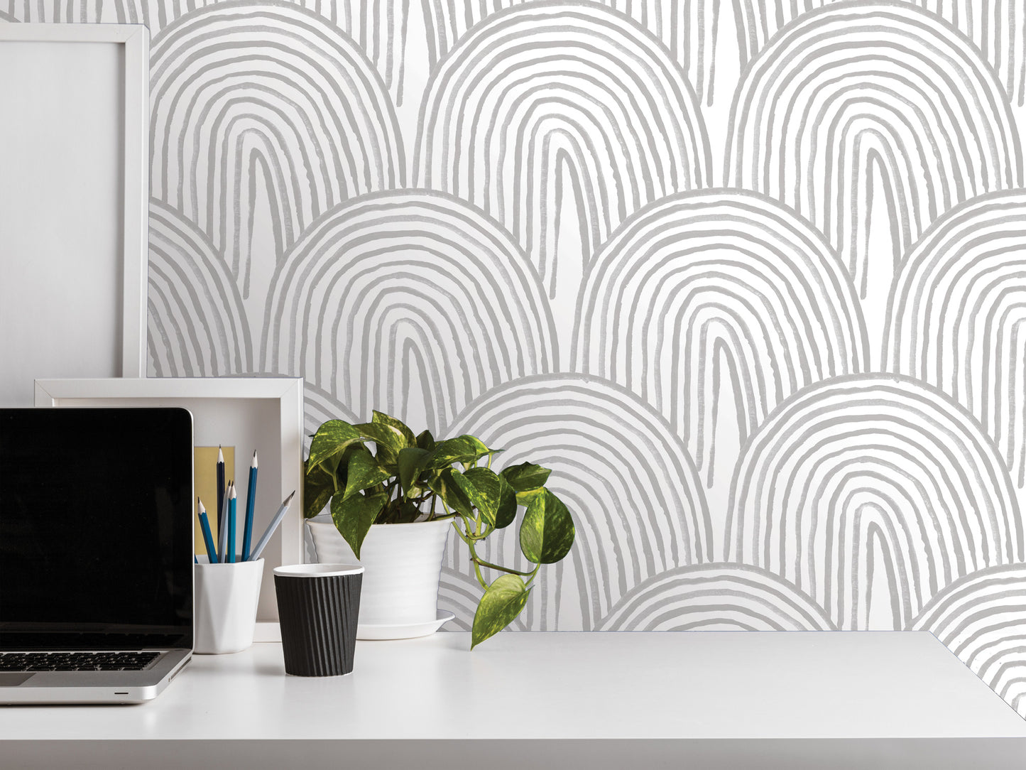 a white desk with various items on it, including a laptop, a plant, a cup, and a framed picture. The background has a repeating pattern of white and gray arches, creating a minimalist and modern aesthetic.