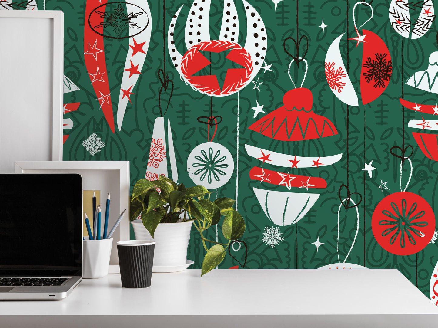 a white desk with a laptop, a cup, and a potted plant. The desk is set against a vibrant green wall with a pattern of Christmas ornaments and stars in red, white, and black.