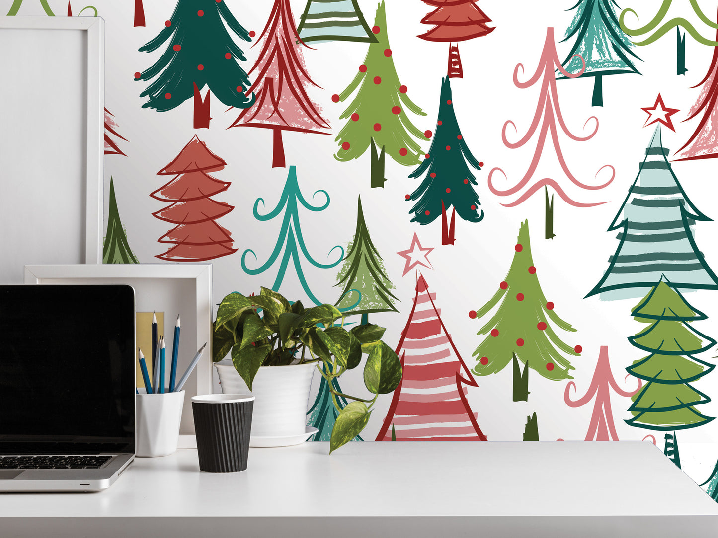 a white desk with a laptop, a cup, and a potted plant. The background behind the desk is decorated with a pattern of colorful Christmas trees in various shades of green, red, and white.