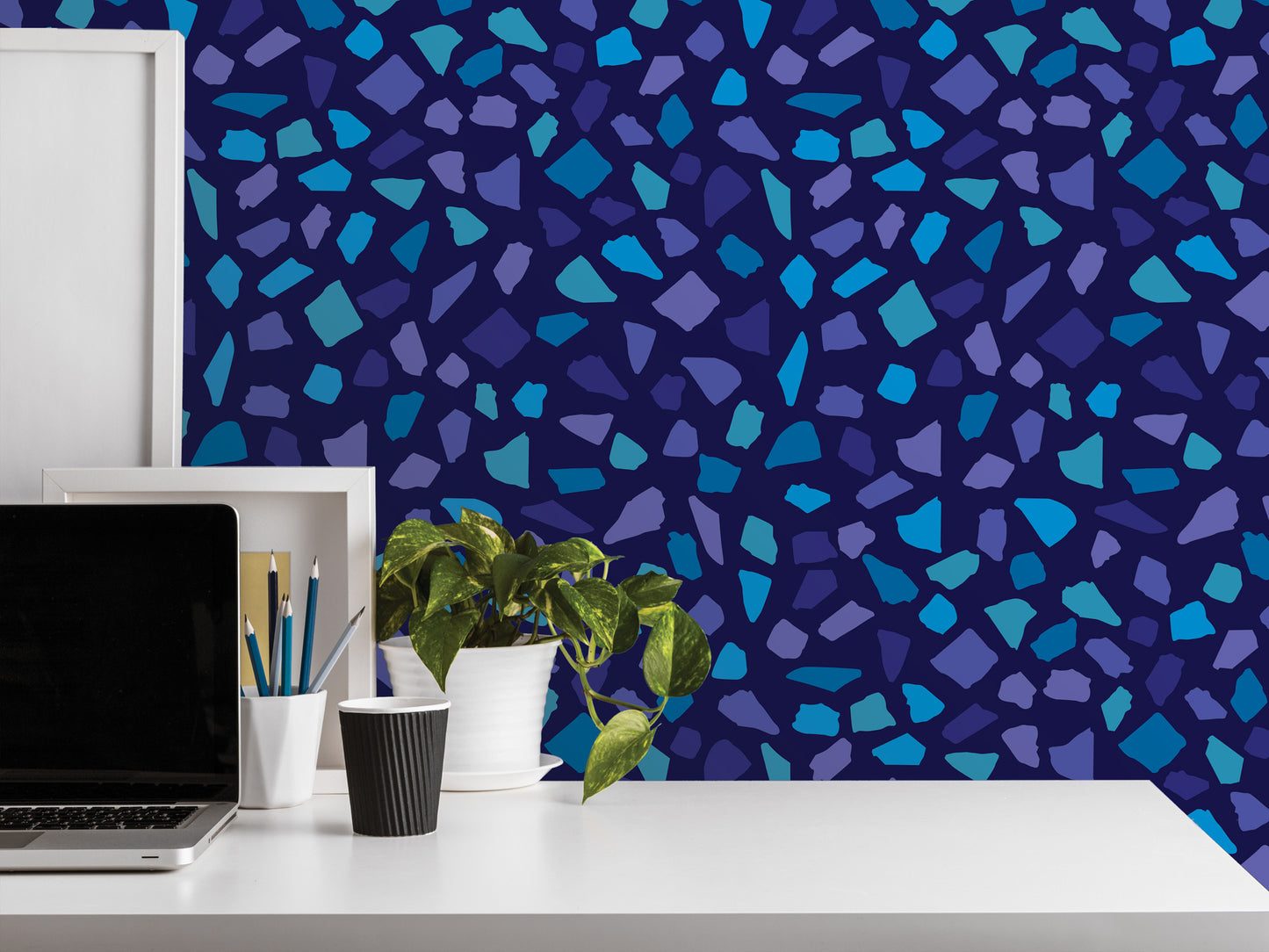 a white desk with a laptop, a plant, and a cup, set against a vibrant blue and purple mosaic patterned wall.