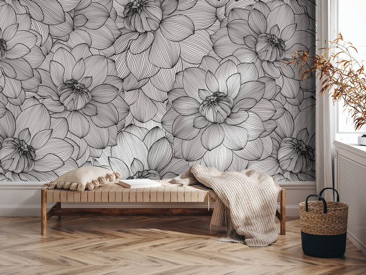 a modern and minimalist interior design with a large wallpapered wall featuring a repeating pattern of black and white flowers. In the foreground, there is a wooden bench with a beige blanket draped over it, and a woven basket next to it. The overall color scheme is neutral, with the black and white flowers providing a striking contrast against the white wall.