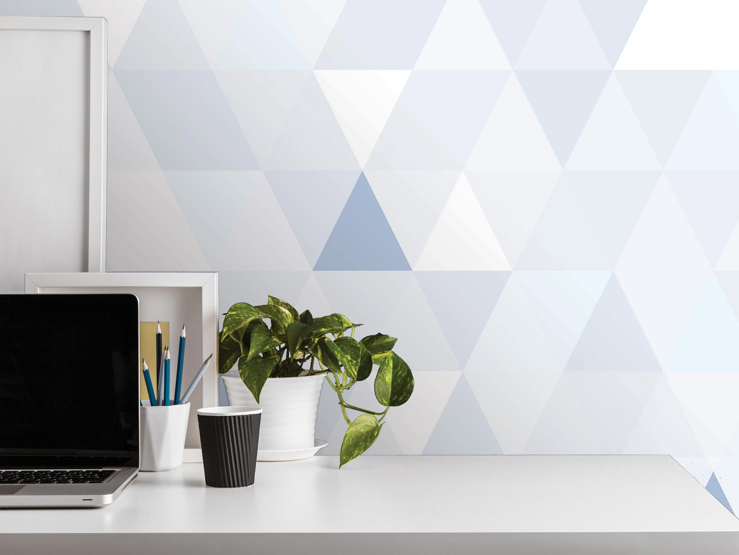 a white desk with a laptop, a black cup, a white vase with a plant, and a white picture frame. The background has a geometric pattern in shades of blue and gray.