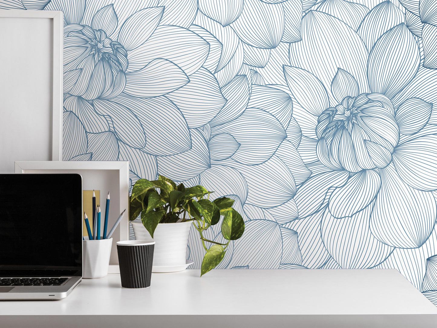 a white desk with a laptop, a black cup, a white vase with a plant, and a framed picture. The background has a blue and white floral pattern wallpaper.