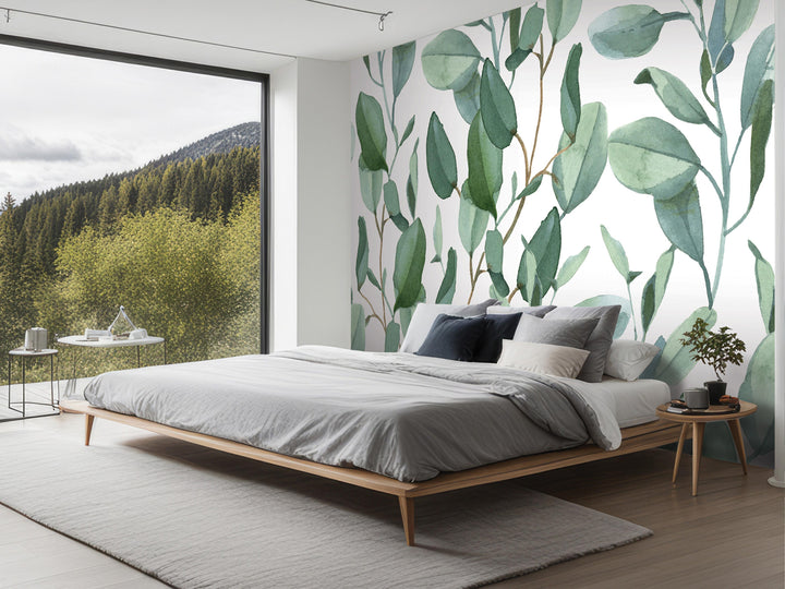 a modern bedroom with a large window overlooking a forested area. The room features a wooden bed frame, a gray comforter, and a green plant on a small table. The walls are adorned with a large mural of green leaves, creating a serene and natural atmosphere in the space.