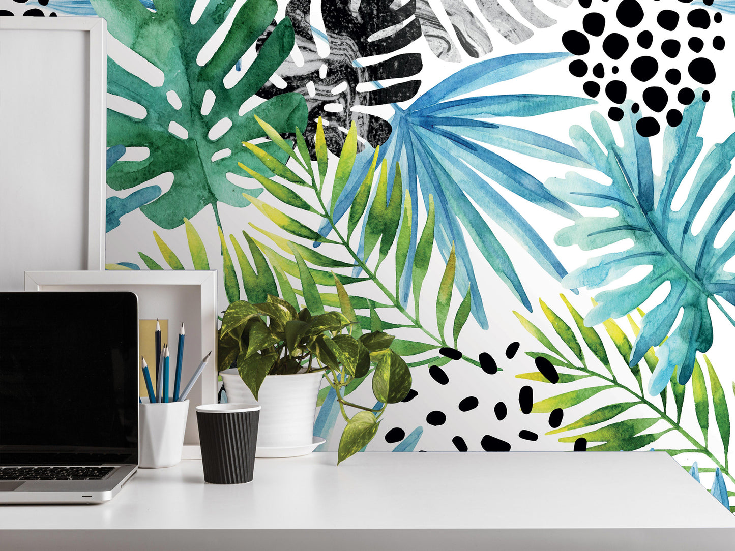 a white desk with a laptop, a plant, and a cup, set against a vibrant wallpapered wall with a tropical leaf pattern.