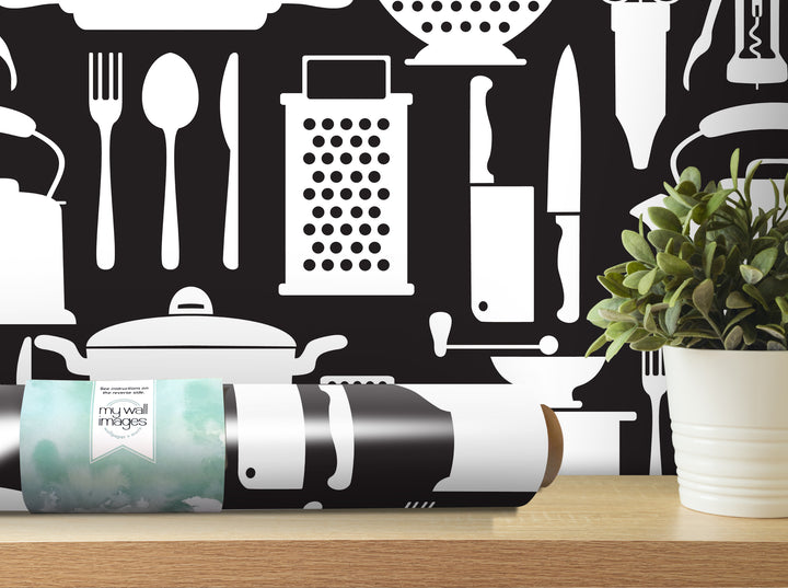 a black and white wallpaper with various kitchen utensils and appliances, such as a knife, a grater, a pot, and a teapot, as well as a potted plant. The wallpaper is displayed on a wall, and there is a roll of wallpaper paper on a table in front of it.