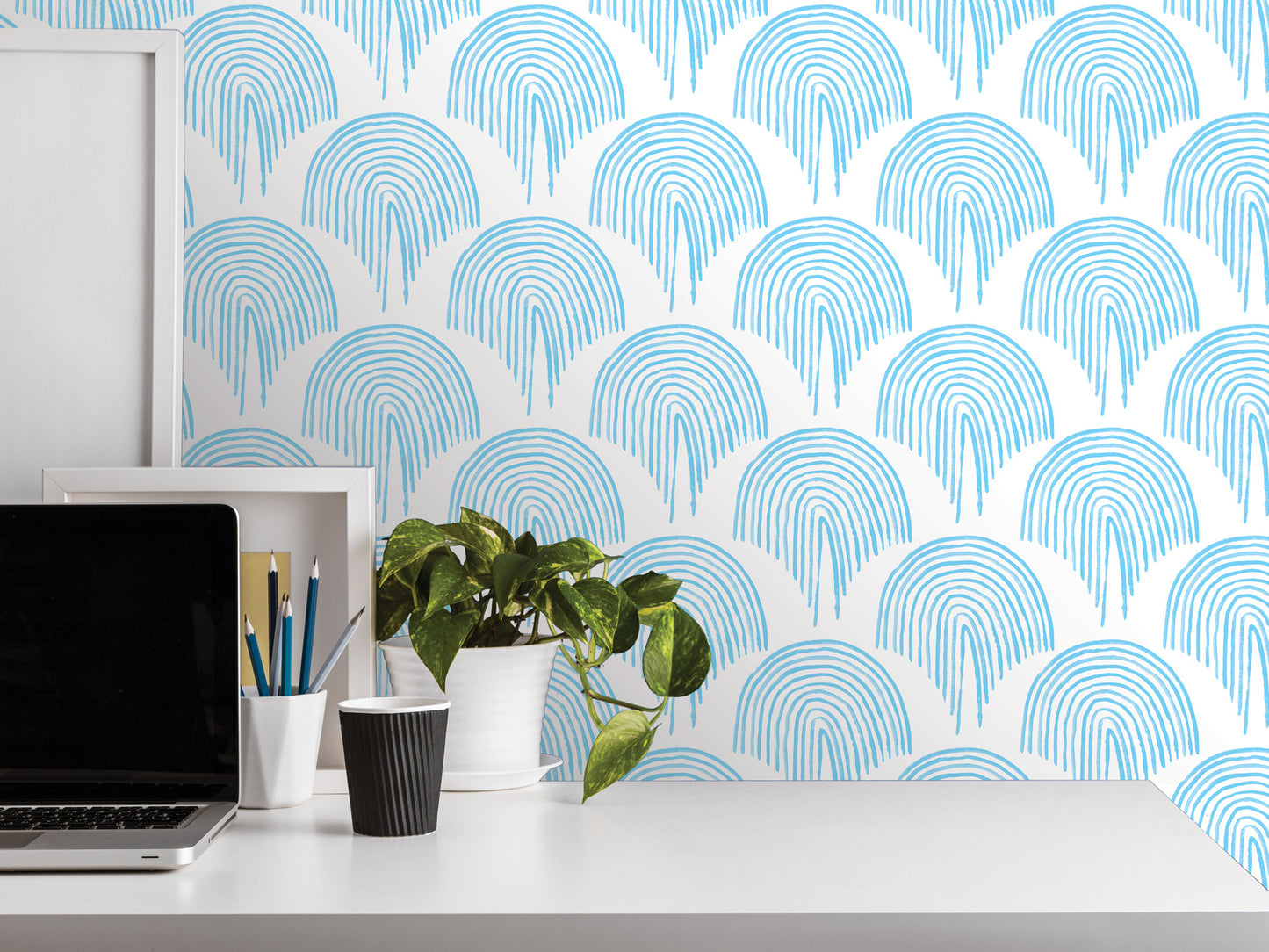 a white desk with a laptop, a black cup, a white vase with a plant, and a framed picture. The background has a blue and white patterned wallpaper with a repeating rainbow design.