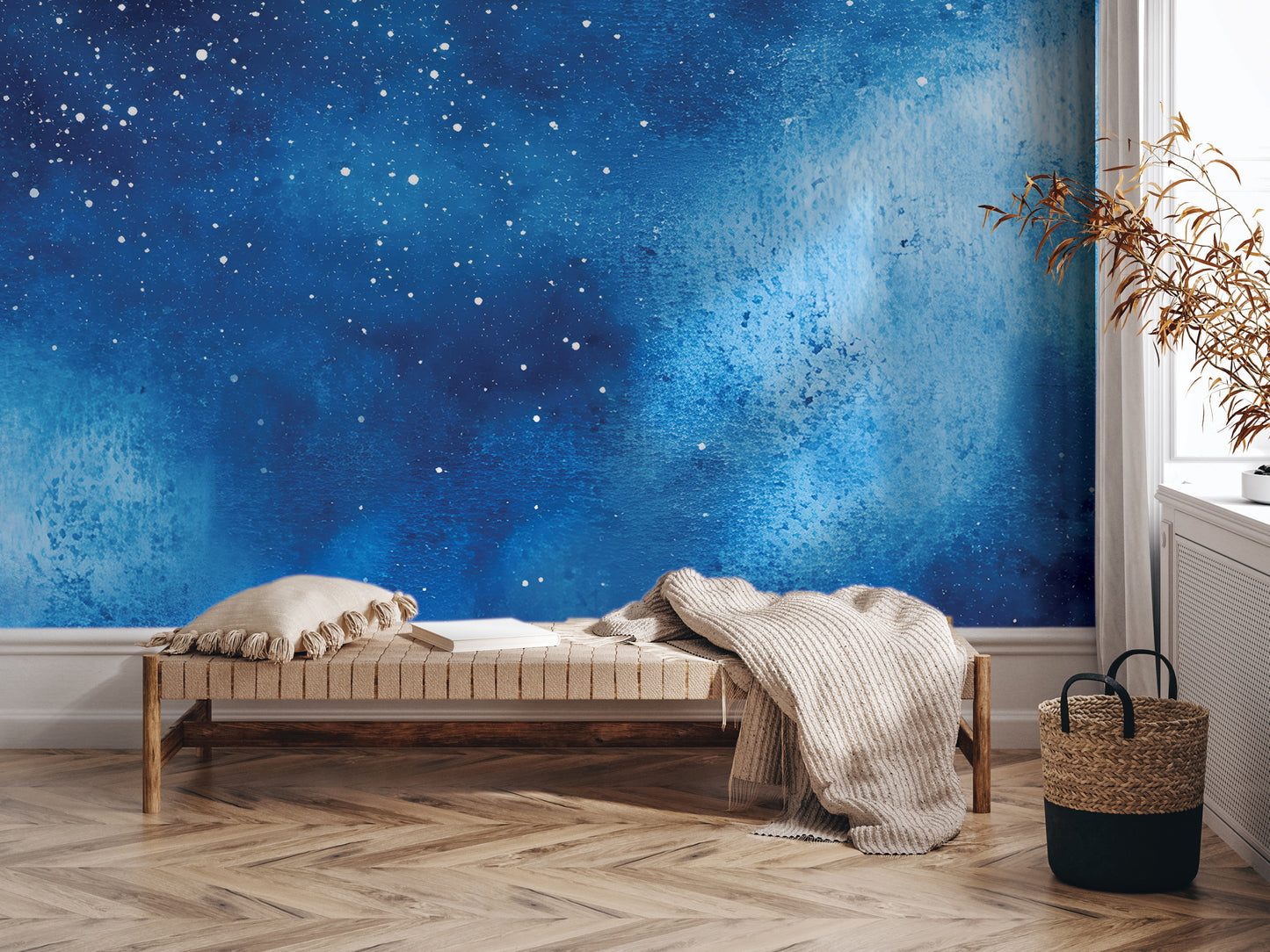 a cozy and inviting living space with a large blue wall mural depicting a starry night sky. In the foreground, there is a wooden bench with a white pillow and a woven basket, creating a comfortable seating area. The room also includes a window and a plant, adding to the overall ambiance of the space.
