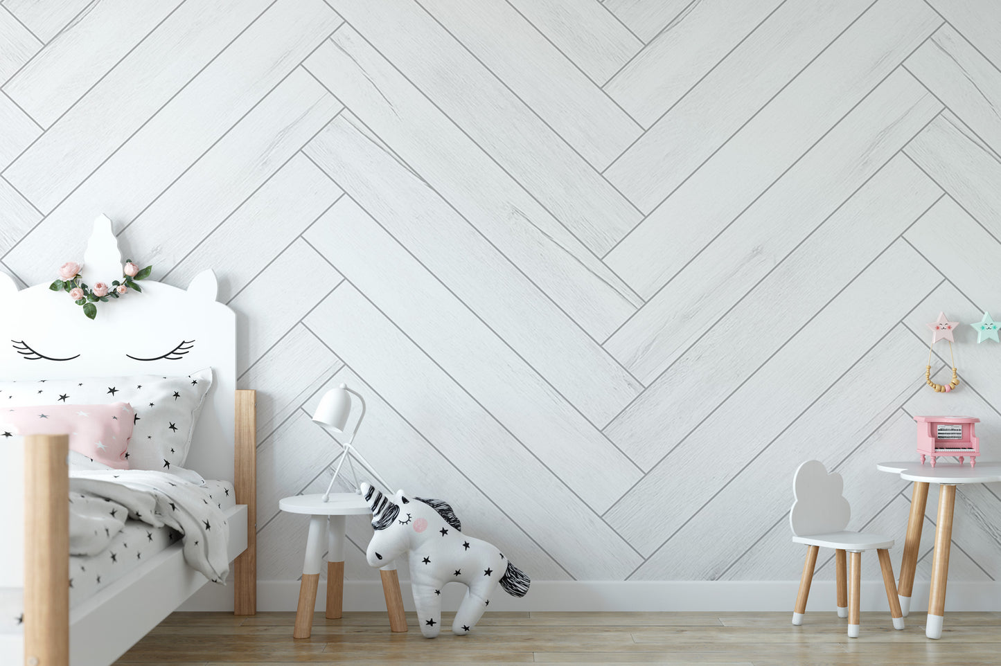 A child's bedroom with a white bed, a small table, and a chair. The walls are covered in a white patterned wallpaper, and there are some decorative elements such as a stuffed animal and a pink toy.