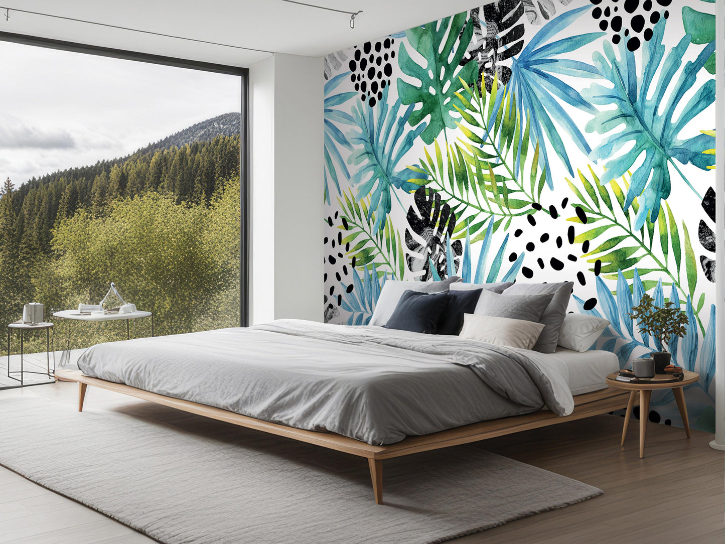 a modern bedroom with a large window overlooking a forested area. The room features a wooden bed frame, a gray comforter, and a wooden nightstand with a plant. The wall behind the bed is decorated with a vibrant tropical wallpaper pattern in shades of green, blue, and white.
