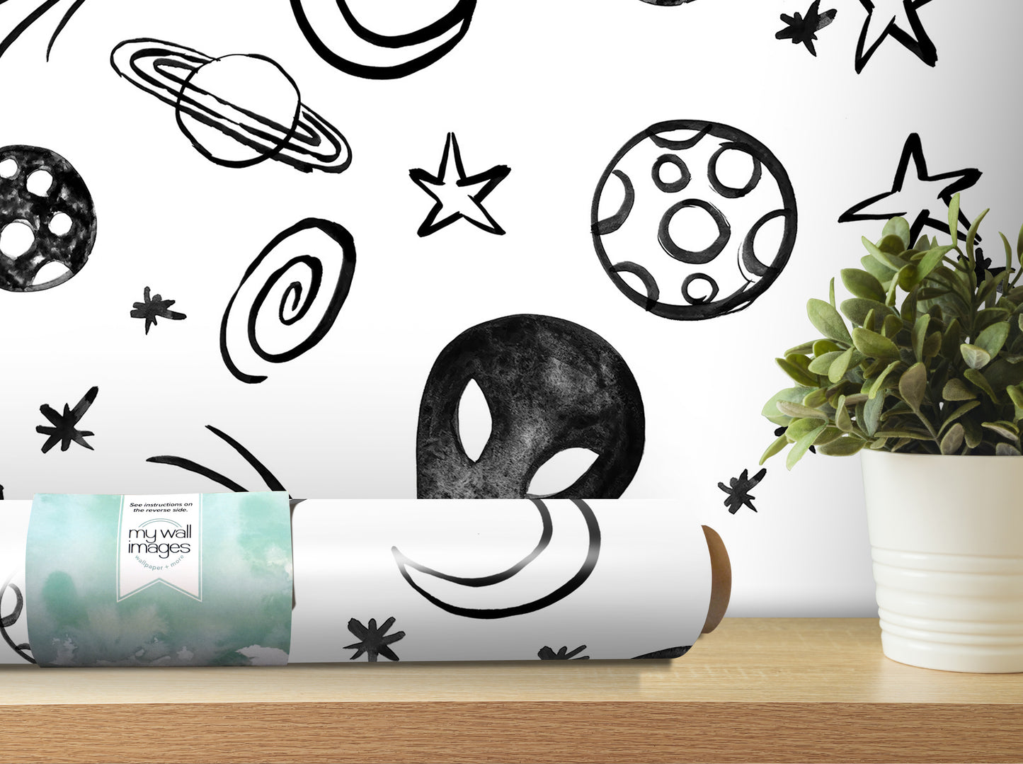 a wall with a black and white space-themed wallpaper, including various celestial objects such as planets, stars, and an alien head. In the foreground, there is a potted plant on a shelf, and a roll of wallpaper is placed on a surface next to it.