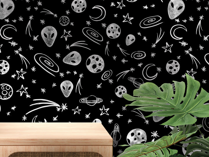 a black and white pattern of various celestial objects, including planets, stars, and alien faces, against a black background. There is a wooden table in the foreground, and a plant is visible in the background.