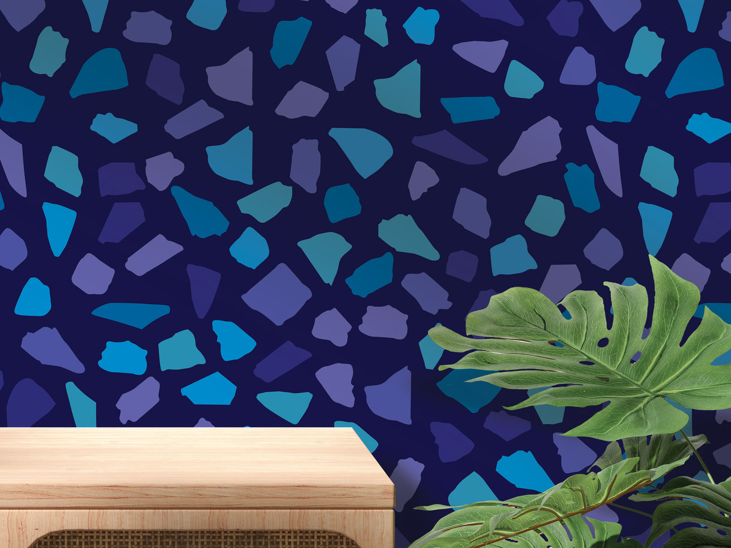 a dark blue wall with a pattern of small blue and purple tiles or stones, creating a visually striking and abstract design. In the foreground, there is a wooden table or shelf, and a large green leafy plant or fern is visible to the right side of the image.