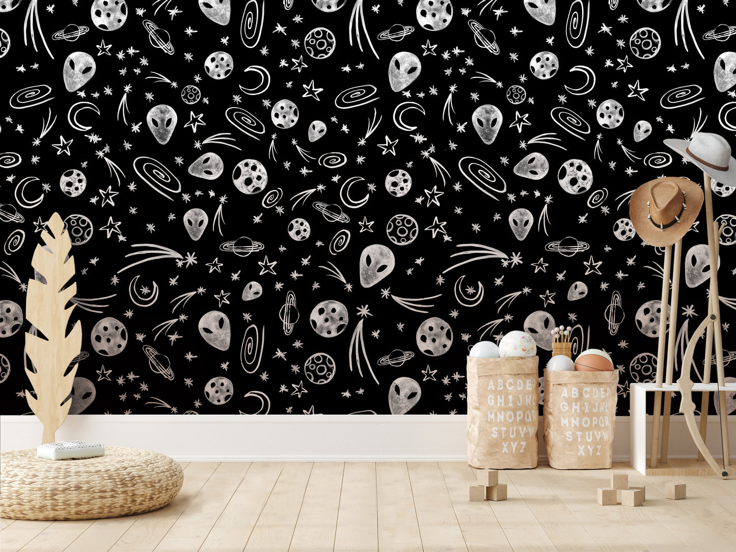 a black and white wall with a pattern of stars, planets, and other celestial objects. In the foreground, there are two wooden stools and a woven basket, and a wooden hat stand. The wall also has a few small objects, such as a small ball and a small plant.