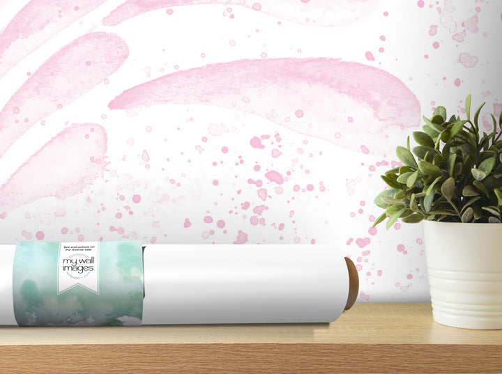 a white wall with a pink watercolor-like pattern, a potted plant, and a roll of wallpaper on a wooden shelf.