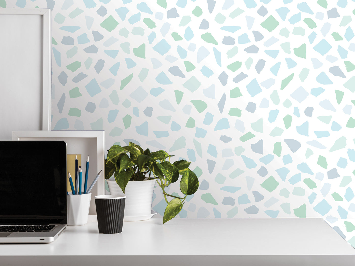 a white desk with a laptop, a black cup, a white vase with a plant, and a white picture frame. The background has a mosaic-like pattern in shades of blue and green.