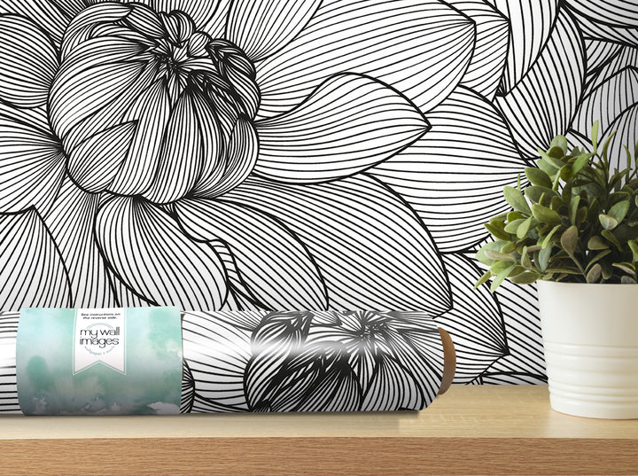 a black and white floral pattern on a wall, with a potted plant and a roll of wallpaper on a shelf in the foreground.