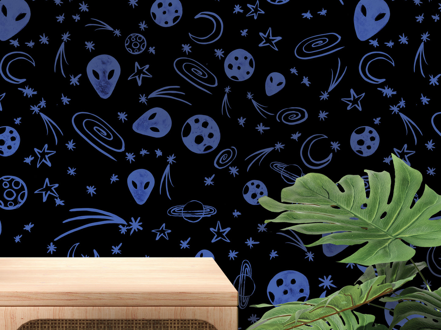 a wall with a pattern of blue alien and celestial objects, including planets, stars, and comets, creating a cosmic atmosphere. In the foreground, there is a wooden table, and a large green plant is visible to the right side of the image.