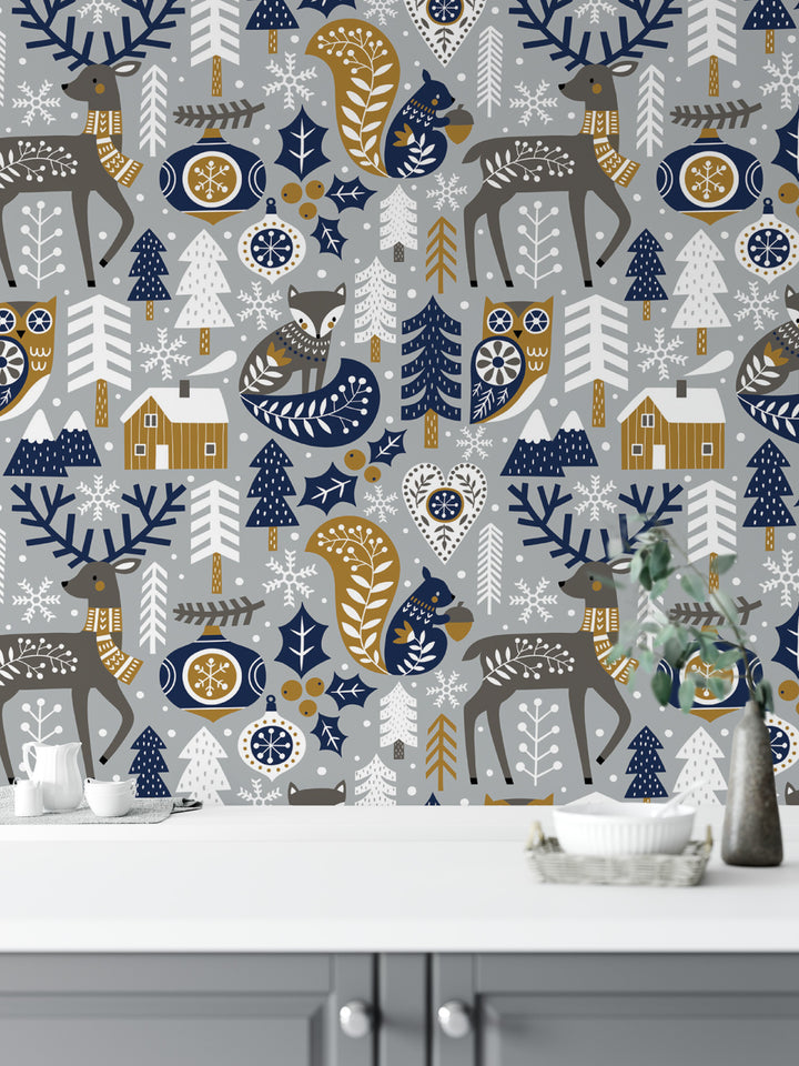 a wallpapered wall with a repeating pattern of various winter animals, such as deer, foxes, and owls, along with snowflakes and pine trees. The wallpapered wall is decorated with a mix of blue, white, and gold colors, creating a festive and wintery atmosphere. In the foreground, there is a white table with a vase and a bowl placed on it.