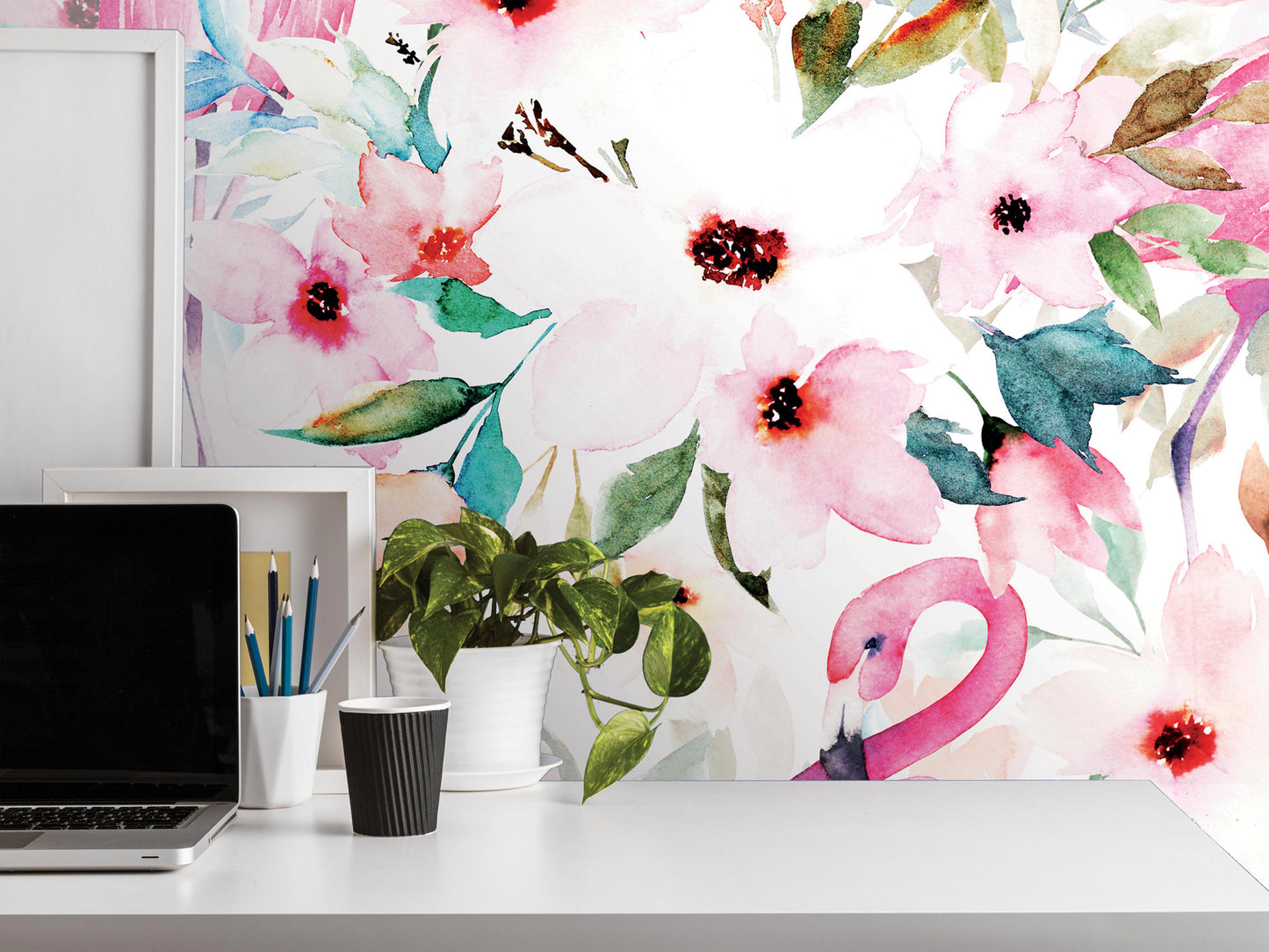 a white desk with a laptop, a cup, and a potted plant. The wall behind the desk has a vibrant floral mural, including pink, green, and white flowers and leaves.