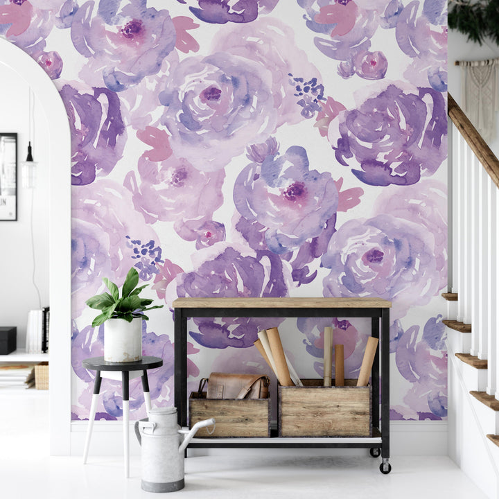 a room with a large wallpapered wall adorned with a floral pattern in shades of purple and pink. In front of the wall, there is a small table with a plant on it, and a wooden crate with various items inside. The room also includes a staircase and a white railing, creating a cozy and inviting atmosphere.