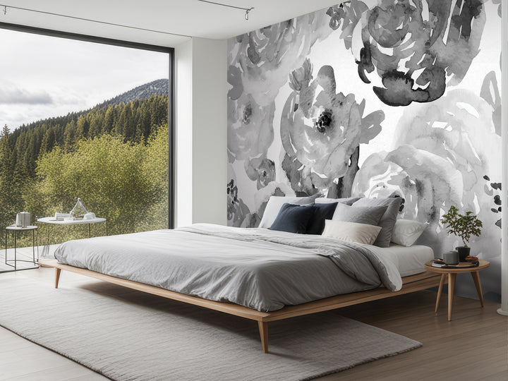 a modern bedroom with a large window overlooking a forested area. The room features a wooden bed frame, a gray comforter, and a wooden nightstand with a plant. The walls are adorned with a large black and white floral mural, and the room has a minimalist and clean design.