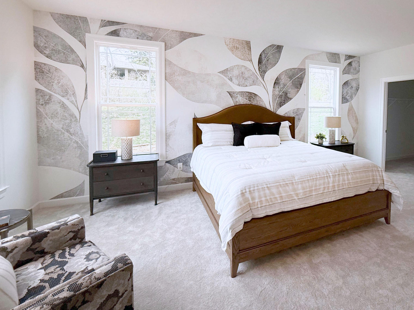 a spacious bedroom with a large bed, a dresser, and a chair. The room has a neutral color scheme with white walls and a gray and white leaf-patterned wallpaper.