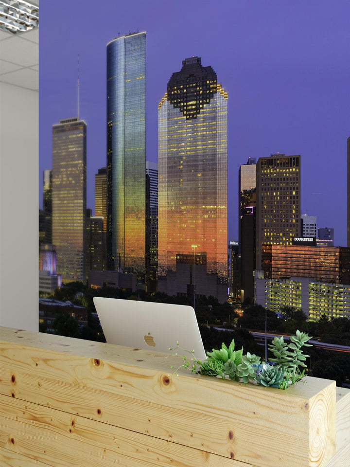 a cityscape with tall buildings at night, including a skyscraper with a distinctive curved shape. In the foreground, there is a wooden desk with a laptop and a potted plant on it.