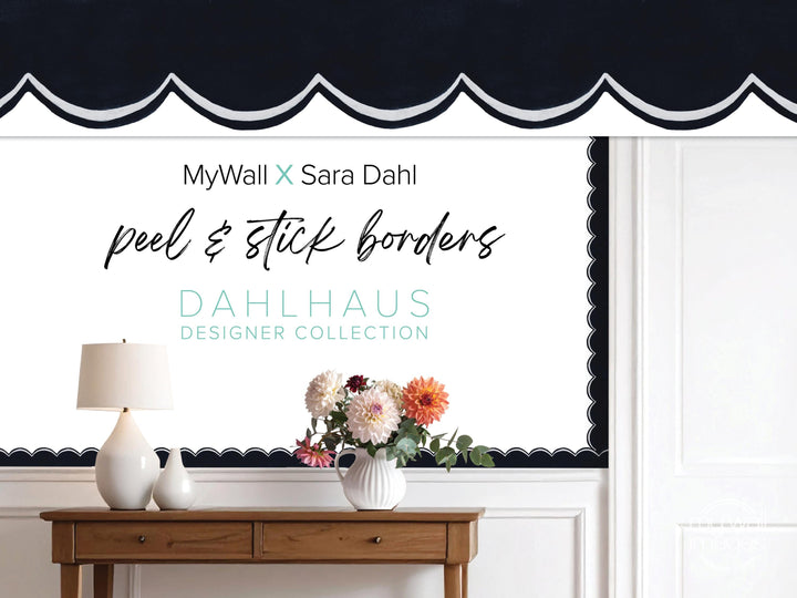 a wooden table with a vase of flowers, a lamp, and a black and white patterned curtain. The curtain has a scalloped edge and the text &quot;MyWall x Sara Dahl peel & stick borders&quot; is displayed on it.