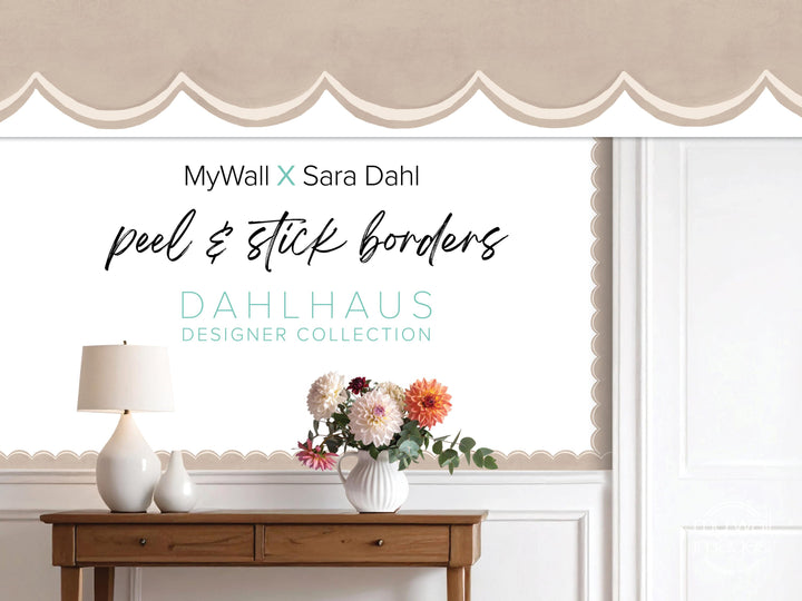 a wooden table with a vase of flowers, a lamp, and a sign for MyWall X Sara Dahl peel and stick borders.