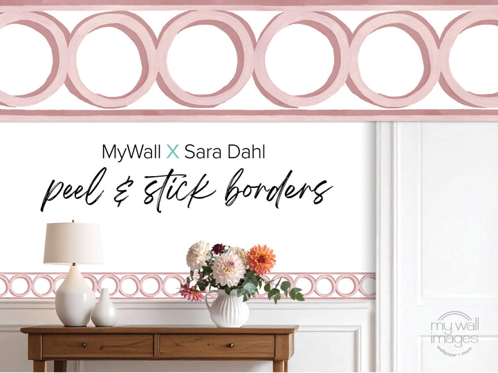 a wooden table with a vase of flowers, a lamp, and a pink border with circular shapes. The text above the table reads &quot;MyWall X Sara Dahl peel & stick borders&quot;.