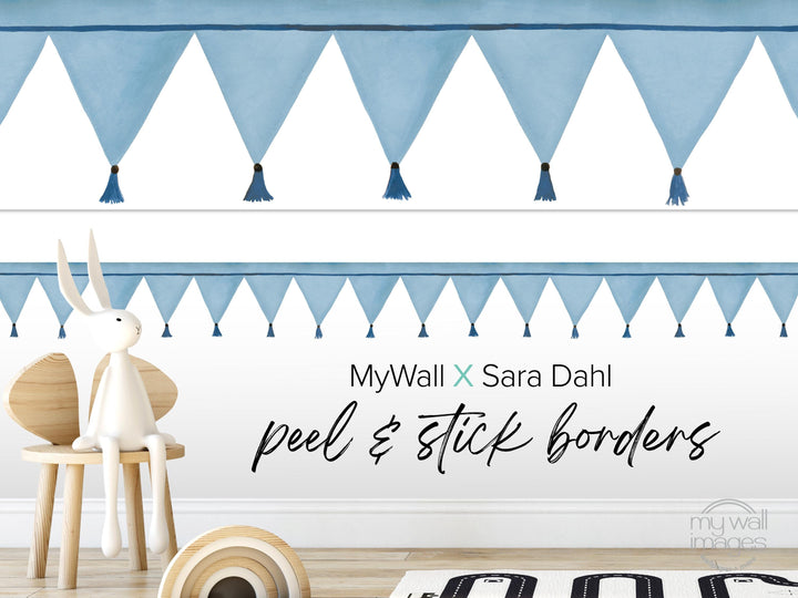 a white rabbit sitting on a wooden chair, with a wall decorated with blue triangular banners and tassels. The wall also has text that reads &quot;MyWall x Sara Dahl peel & stick borders&quot;.
